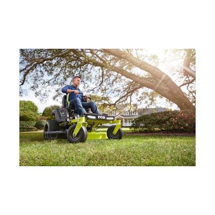 En person kör en grön Ryobi åkgräsklippare på en gräsmatta med träd och buskar i bakgrunden.