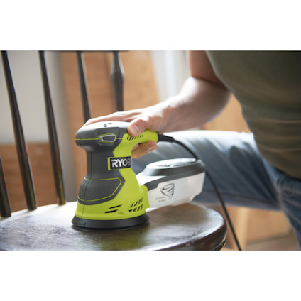 Person använder en grön och svart Ryobi-slipsmaskin på en stol.