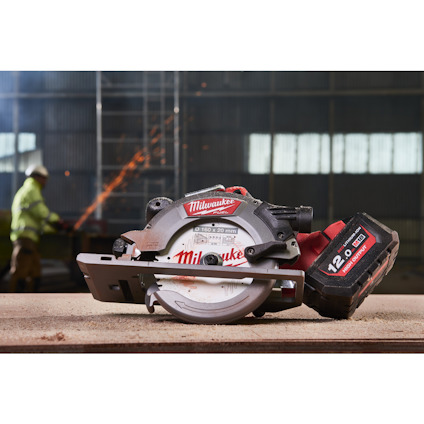 Cirkelsåg från Milwaukee med en stor batterienhet på ett bord, i bakgrunden en arbetare och gnistor.