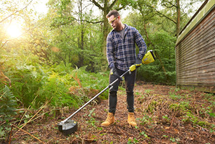 En person trimmar buskar i en skog med en grön och svart trimmer, iklädd rutskjorta och arbetsstövlar.