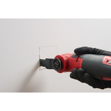 Hand håller ett oscillerande verktyg och skär ett fyrkantigt hål i en vit vägg.