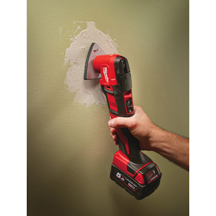 En person använder ett rött, batteridrivet verktyg med osynliga varumärkesdetaljer mot en vägg med gips.