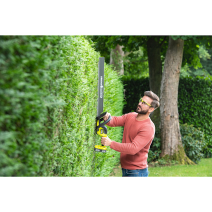 Man in orange sweater using a cordless hedge trimmer on a green bush, wearing safety glasses in a garden setting.