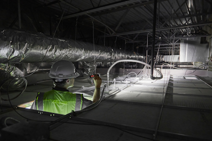Person i skyddshjälm och väst inspekterar ventilationssystem i mörkt industriellt takutrymme.