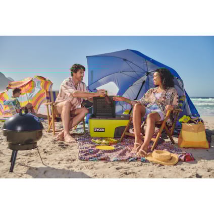 En man och en kvinna sitter på en sandstrand, omgivna av färgglada parasoller och en grill nära en RYOBI kylbox.