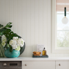 Tvättstuga med vägg av vit pärlspontpanel, fönster med matchande foder, en turkos vas med vita blommor och gröna blad på bänken. Lampa i fönstret.