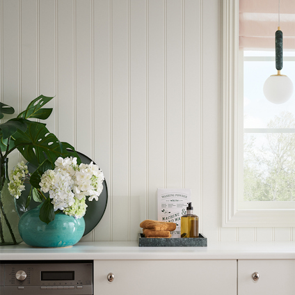 Tvättstuga med vägg av vit pärlspontpanel, fönster med matchande foder, en turkos vas med vita blommor och gröna blad på bänken. Lampa i fönstret.