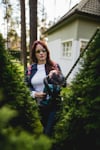 Kvinna med hörselskydd klipper buskar med en batteridriven häcksax nära ett hus.