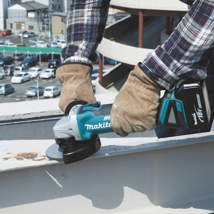 Person som använder en Makita vinkelslip på en metallkant utomhus, med bilar och byggnader i bakgrunden.