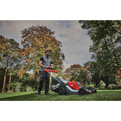 Man pushing a lawnmower on grass, surrounded by trees with autumn foliage.