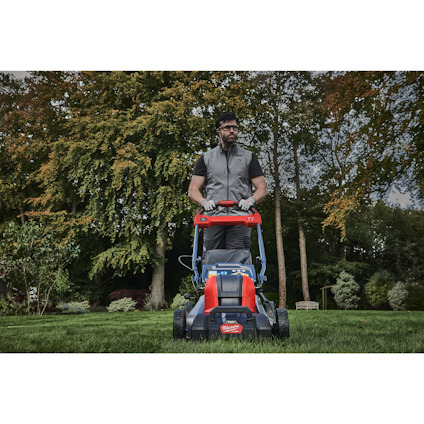 Man mowing lawn with a red and blue lawnmower in a garden setting with trees in the background.