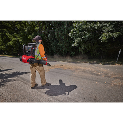Person med lövblåsare står på trottoar med träd i bakgrunden.