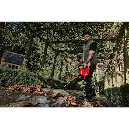 Person i mörka kläder använder en röd lövblås under en grön pergola, omgiven av buskar och löv.