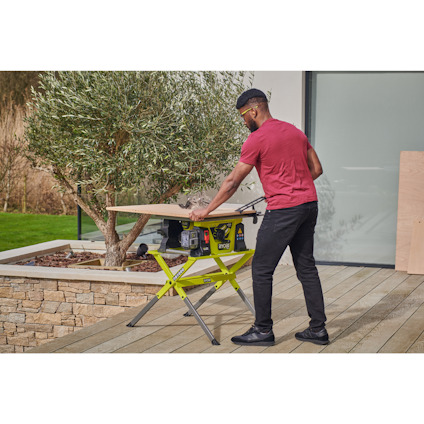 Person i röd tröja använder en bordsåg som står på ett anpassat stativ på en terrass bredvid ett olivträd och en glasvägg. Samtliga produkter är av märktet Ryobi