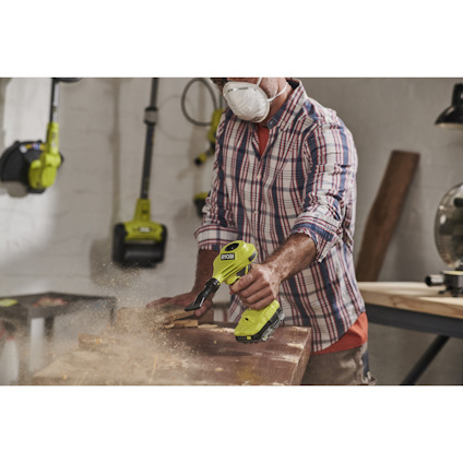 Person dammsuger bort sågspån i verkstaden med en volympump från Ryobi, bär skyddsglasögon och ansiktsmask.