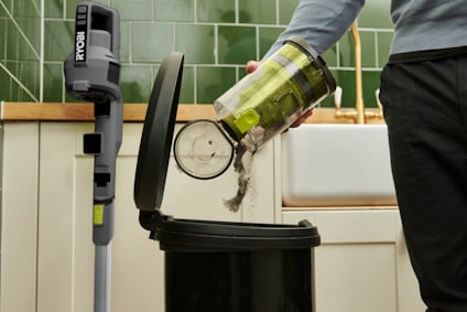 En person tömmer en dammsugarbehållare i en soptunna. Skaftdammsugaren av märket RYOBI står mot en köksbänk.