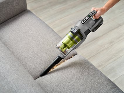 Hand håller en Ryobi skaftdammsugare som suger damm från en grå soffa.