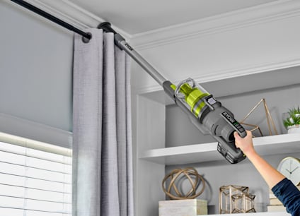 Person som använder en grå RYOBI skaftdammsugare för att rengöra gardinstång nära taket.