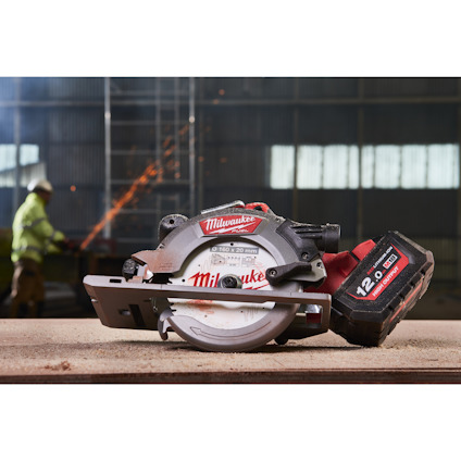 Cirkelsåg från Milwaukee med batteri bredvid, i ett industriellt miljö med en svetsande arbetare i bakgrunden.