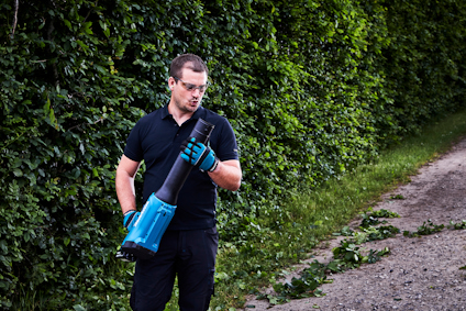 Person i skyddsglasögon använder en lövblås vid en grusväg och häck.