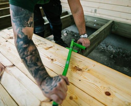 En person använder en skruvdragare för att fästa en träskruv med försänkt huvud i träplankor utomhus under en klarblå himmel.