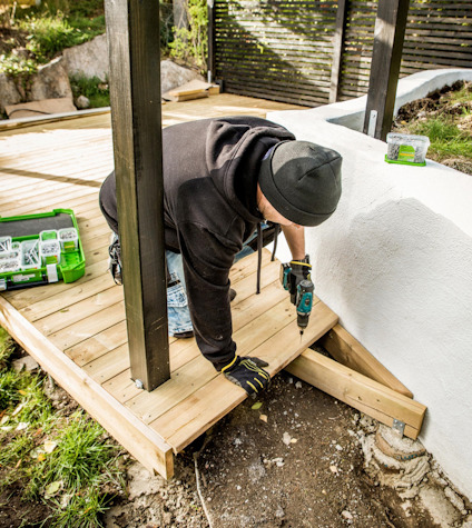 Person i mörka kläder bygger en träaltan, använder skruvdragare. Grön vegetation i bakgrunden.