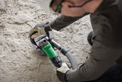 Person använder ett elverktyg för att slipa en betongyta. Verktyget har ett grönt handtag och en dammsugarslang ansluten.