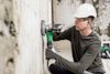En person i hjälm och hörselskydd använder en grön elektrisk slipmaskin på en vägg.