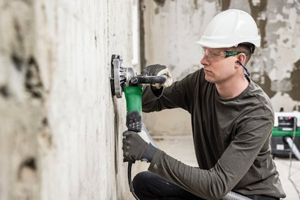 En person i hjälm och hörselskydd använder en grön elektrisk slipmaskin på en vägg.