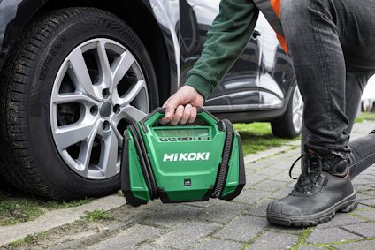 Person kneeling by car wheel, holding a green Hikoki tool case.