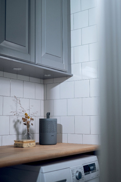 Gråa köksskåp, vita kakelväggar, träbänk med en svart apparat och liten vas med torkad växt. Tvättmaskin under bänken.