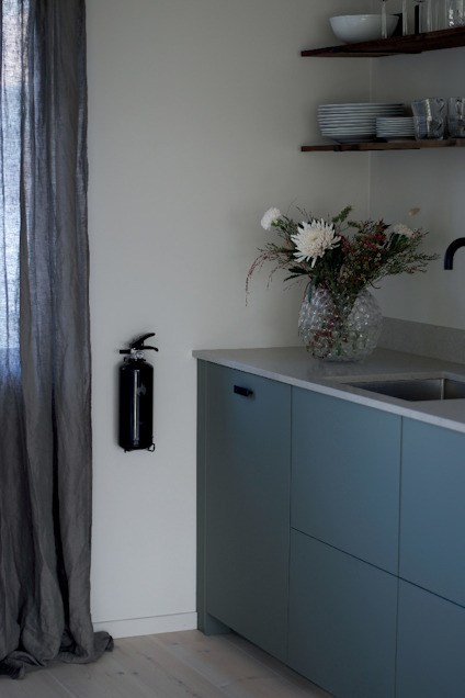 Grått kök med blå skåp, beige vägg, brandfilt, diskbänk, hyllor med tallrikar och ett blommarrangemang i glasvas.