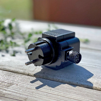 Elektrisk adapter med två kontaktstift och en skruvkoppling ligger på en träyta. Bakgrund med gröna växter.